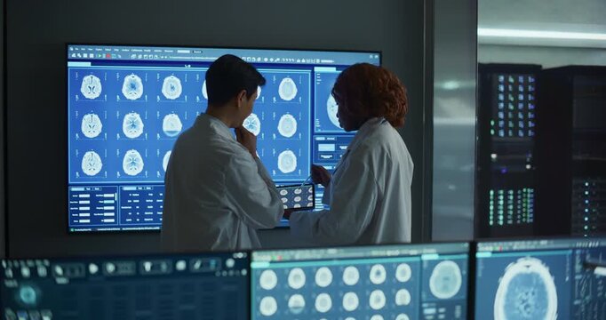 Black Female Neurologist and Asian Male Doctor Meeting and Talking, Reviewing MRI Brain Scans on a Large Display, Hospital Laboratory. Neuroscience Research and Disease Treatment. Backview
