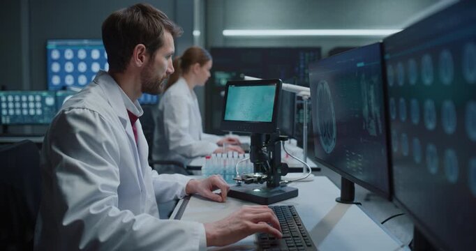 Male Scientist Inspects Biological Samples With a Digital Microscope While checking MRI Brain Scans Displayed on Monitors. Research Team Pursues Neurological Drug Innovation in Hospital Lab.