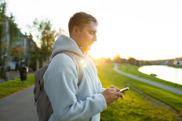 man relaxing on pathway in park at sunset with backpack and phone in hand side view. man wearing fleece jacket and backpack walking on concrete pathway with grass and trees in background. © Shi 
