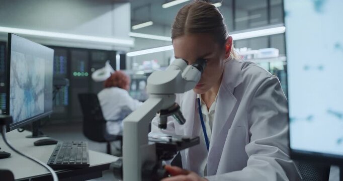 Portrait of Caucasian Female Scientist Looks Into Digital Microscope to Analyze Molecular Samples Beside Screens Displaying Scan Data as Colleagues Advance Pharmaceutical Disease Research in Lab