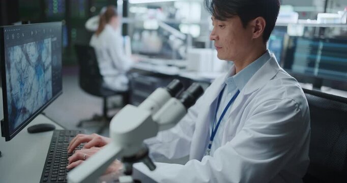 Portrait of Asian Male Scientist Looking Under Microscope Specimens Then Studies Cellular Data on Computer Monitor in Research Hospital Laboratory. Drug Development for Disease. Slow Motion