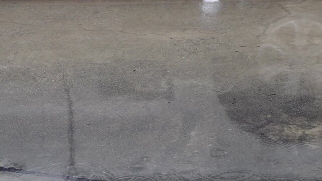raindrops creating ripples on the water surface. Dark grey concrete road texture with puddles. Moody urban background for weather or autumn scenes.