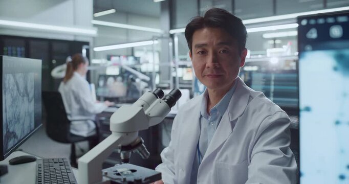 Portrait of Asian Male Scientist Seated Beside Digital Microscope in Advanced Medical Research Hospital Lab, Looking at Camera While Colleagues Studies Cellular Data on Screens for Drug Development