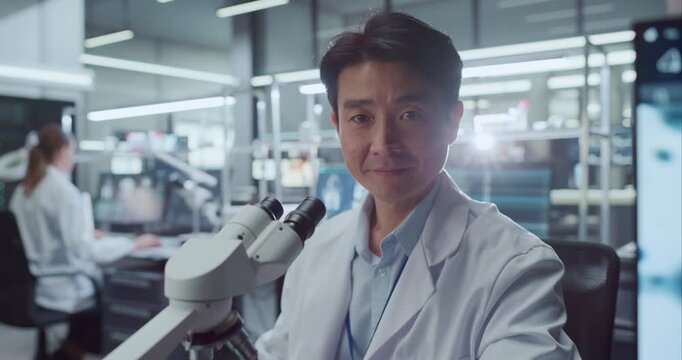 Portrait of Asian Male Scientist Seated Beside Digital Microscope in Advanced Medical Research Hospital Lab, Looking at Camera While Colleagues Studies Cellular Data on Screens for Drug Development