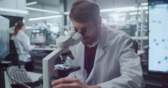Portrait of Caucasian Male Scientist Looking Through Digital Microscope to Examine Biological Samples in High-tech Medical Research Hospital Lab, Female Colleague Studies Data for Disease Treatment