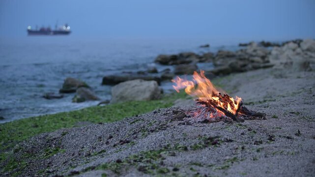 Bonfire burning on a rocky beach at dusk with cargo ship on the horizon