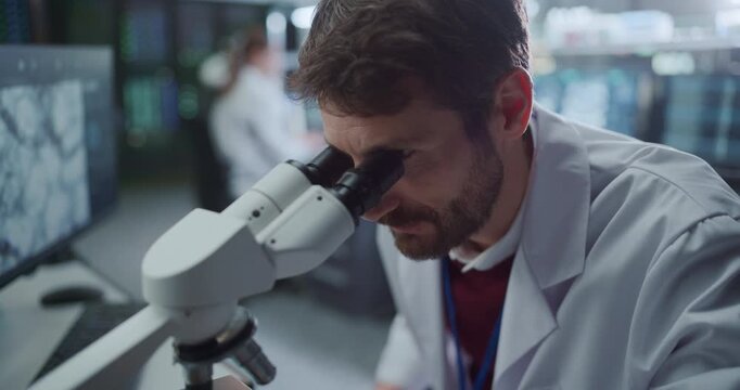 Portrait of Male Scientist Looking Under Microscope Inspecting Biological Samples in Medical Hospital Laboratory While Colleagues Reviews Molecular Data Screens for Drug Development. Slow Motion