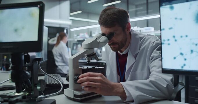 Portrait of Male Scientist Looking Under Digital Microscope to Examine Biological Specimens in Medical Research Hospital, as Female Colleague Studies Molecular Data on Screens for Disease Treatment