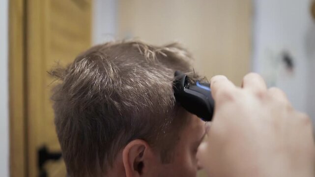 A man receives a haircut in a professional barber shop, viewed from the side. The scene features hair clippers and a mirror, highlighting the grooming process. The setting is well-lit.