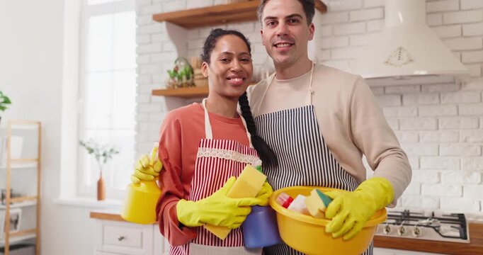 Couple cleaning home kitchen with teamwork. Smiling partners in aprons wear gloves and hold a bucket with sponges, posing in a bright tidy kitchen. Concept shows teamwork and happy housekeeping.
