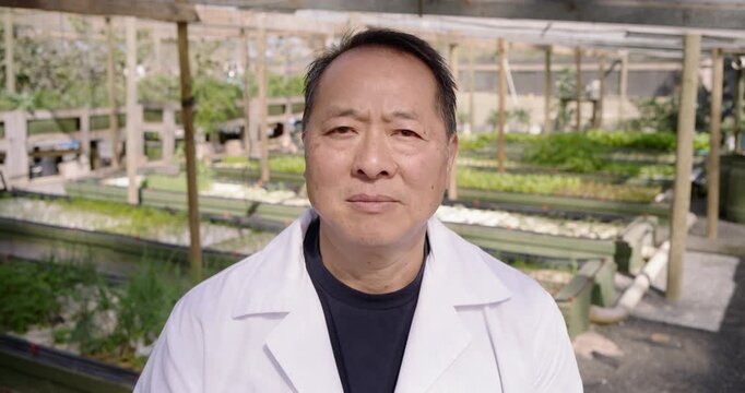 Senior Asian man in nursery lab coat, softening and smiling, responding to camera near troughs