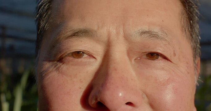 Senior Asian man blinking eyes, squinting, and smiling while adjusting to bright sunlight in garden