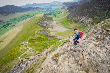 Fototapeta premium A female hiker descending the rocky ridge of High Snab Bank, ridgeline, from the summit of Robinson with Newlands in the distance in the English Lake District, UK.