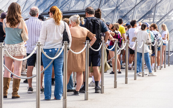 People stand in line &mdash; alone, as a couple, in a group, with family or friends &mdash; between stainless steel queue poles with grey ropes at the entrance of a tourist site, on a sunny summer day.