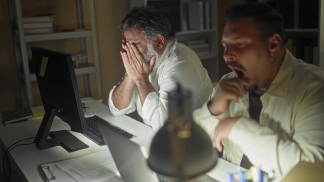 Two men at a desk covering faces with hands by a computer monitor in an office building, one stretching and the other rubbing his eyes; late night fatigue.