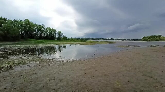 The gently sloping sandy shore of a shallow lake and tall bushes on the opposite shore on a sunny summer day. Matveevskaya Channel, Siberia, Russia.