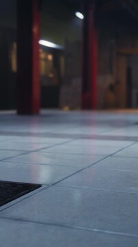 The scene shows tiles on a floor with a light source illuminating the area. Shadows form along the surface, and a vent is visible nearby. The urban space suggests activity.