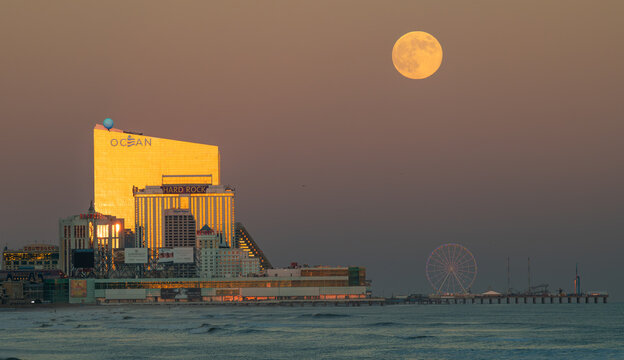 Atlantic City skyline with illuminated casinos, Ocean and Hard Rock hotels reflecting sunset light under a rising full moon above the boardwalk amusement pier and calm Atlantic Ocean at dusk.