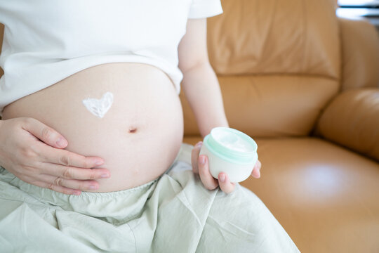 Pregnant woman applying heart shaped stretch mark cream to her belly while sitting on the sofa at home. Preganacy body care routine concept.