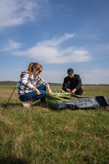 Naklejka premium Young woman and man collaborating to set up a camping tent in a grassy field under a clear blue sky, preparing for an outdoor adventure and leisure activity. Vertical photo