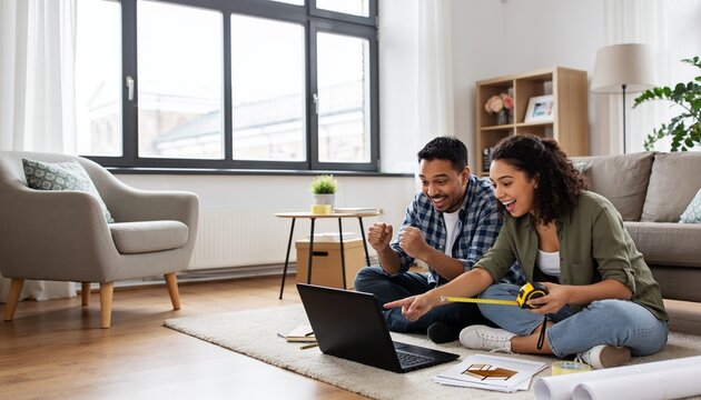 Excited young couple sitting on living room floor with laptop and tape measure, planning home renovation and interior design for new apartment or house moving