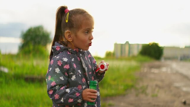 Little girl with hearing aids blowing soap bubbles in slow motion, framed with cinematic anamorphic lenses in a serene outdoor setting at golden hour