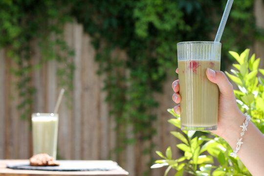 A hand holds a glass with a straw, filled with a light-colored drink like matcha tea. Another similar glass sits on the table behind it.