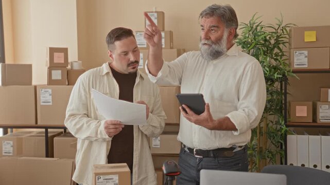Two men hold tablet and papers, one points finger while checking packages and shelves in a building warehouse; teamwork planning.