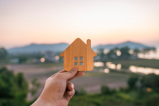 Hand holding a small wooden house icon with sunset nature background. Real estate property, insurance, home mortgage loan, refinance, eco living and family land concept.