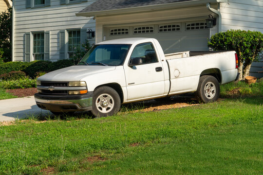 White Chevrolet Silverado pickup truck parked on gravel next to a suburban house with a garage and green lawn, depicting utility, transport, and everyday life in a sunny residential setting.