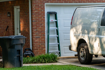 White utility van parked in a sunny residential driveway with a green stepladder leaning against a white garage door, next to a brick house, trash can, and lawn equipment. © Michael