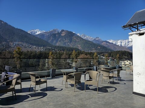 Empty outdoor cafe terrace with wicker chairs and tables overlooking majestic snow-capped Himalayan mountains under a clear blue sky in Manali.