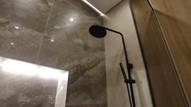 Upward moving view reveals matte black rain showerhead on curved arm beside vertical slide rail, framed by grey marble, warm wood walls, recessed niche, frameless glass divider, modern bathroom