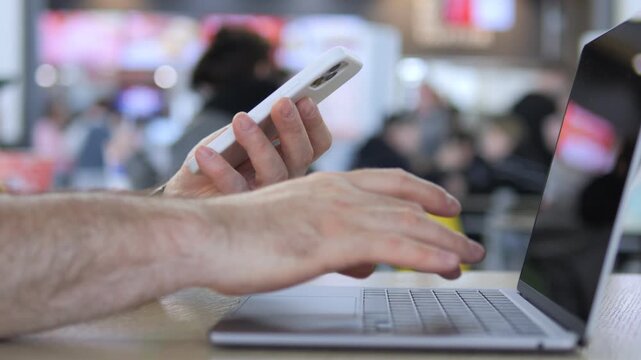 Person multitasking with phone and laptop in public space. Hands show focused activity on devices. Background shows blurred crowd in cafe or lounge area