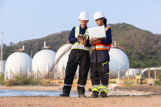 Two engineers safety helmet and reflective vest holding laptop and tablet standing near large spherical storage tank industrial facility with focused expression discussing project details outdoors