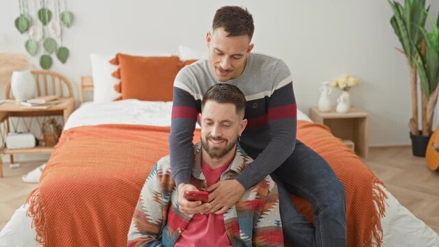 Man couple seated on bed, one man embraces partner with hand on chest while both look at a smartphone in a bedroom building; intimacy.