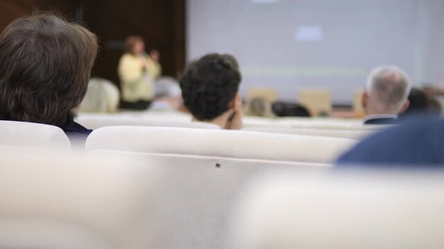 Audience seated in rows faces speaker at conference. People listen attentively, heads turned forward. Focus on back of attendees' heads, blurred background. Ideal for business, education