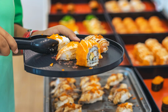 Woman customer choosing various sushi rolls with tongs at Japanese buffet restaurant. Hand of woman using tongs to pick fresh salmon nigiri sushi onto plate from food display tray at Japanese buffet.