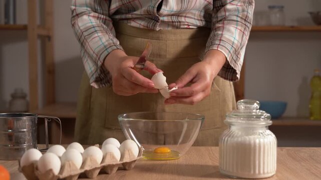 A close-up of a woman's hands cracking an egg with a knife over a clear glass bowl. Homemade food, home cooking