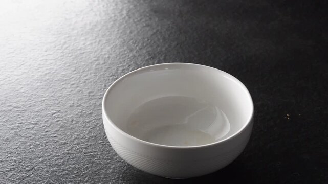 Minimalist and elegant scene showing a single empty white ceramic bowl sitting on a dark textured surface as a light source moves across it, creating shifting highlights and shadows