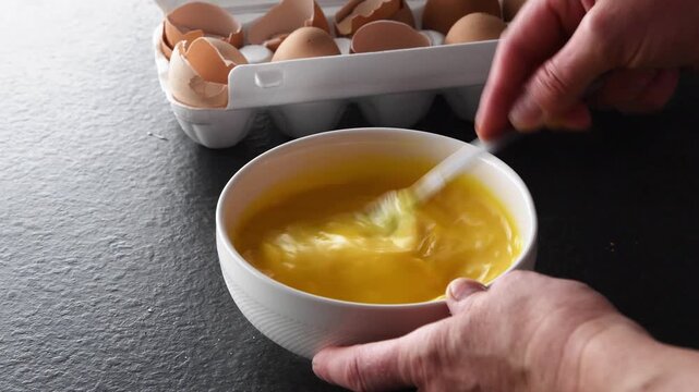 Close up of a person's hands rapidly whisking fresh brown eggs with a fork in a white bowl, preparing a nutritious breakfast recipe on a dark stone countertop in the kitchen