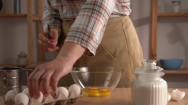 A close-up of a woman's hands cracking an egg with a knife over a clear glass bowl. Homemade food, home cooking.