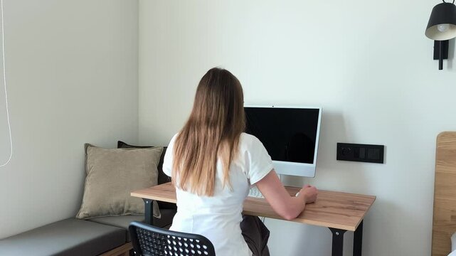 Woman enters her home office and sits down at the computer desk, switching on her monitor. Back view shows her settling into the chair to begin her tasks. A remote workday starts for a