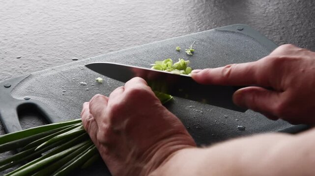 Professional chef preparing a healthy meal by chopping fresh green onions with a sharp kitchen knife on a dark cutting board, a close up view of the skilled food preparation process
