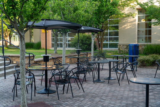 Modern outdoor cafe patio with black tables, chairs, and solar-powered umbrellas on a paved brick courtyard, surrounded by lush green trees and a contemporary building.