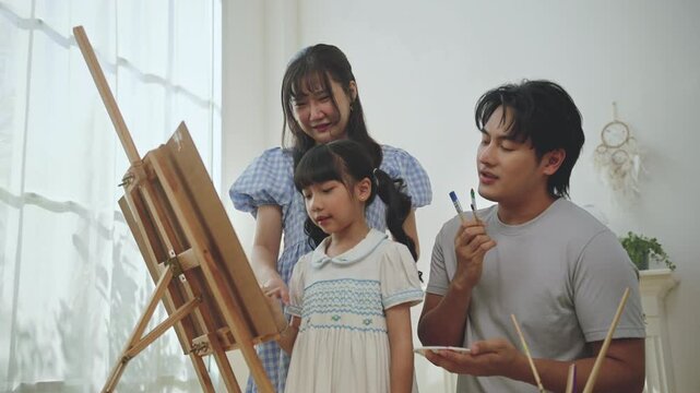 Asian family enjoying painting together at home. father, mother, and young daughter doing creative art activity with paint and canvas in a bright living room, creativity, and learning through play.