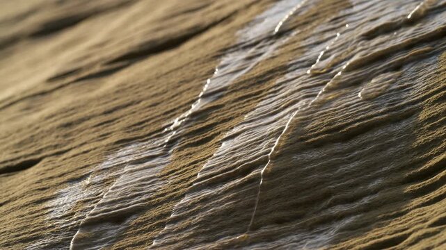 Diagonal close view of rippled beach sand with thin seawater films catching warm golden side light, showing textured ridges, glossy wet channels, and calm coastal detail