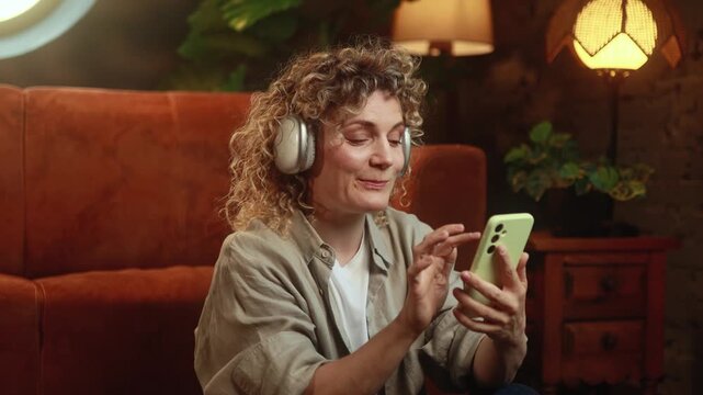 Horizontal video of relaxed curly haired woman listening to music in headphones while scrolling smartphone at home. High quality 4k footage