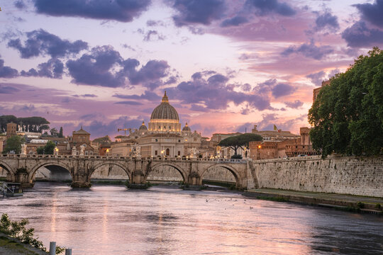 Beautiful illuminated view of Rome, Italy with Saint Peters Basiica, historic architecture, bridges and Tiber River under evening sky. 