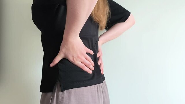 Woman touches her lower back with both hands reacting to sudden pain. She arches slightly while feeling discomfort along her spine. Concept of lumbar strain, posture problems and chronic